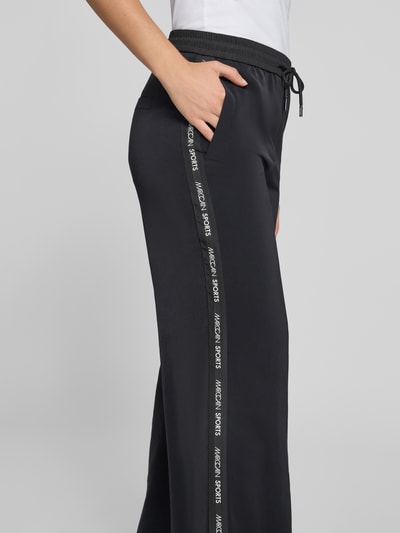 Frauenmodel trägt Schlupfhose mit seitlichen Label-Details und elastischem Bund von Marc Cain, Relaxed-Fit, schwarze Stoffstruktur, Eingrifftaschen, Tunnelzug, Detailaufnahme Seitansicht.