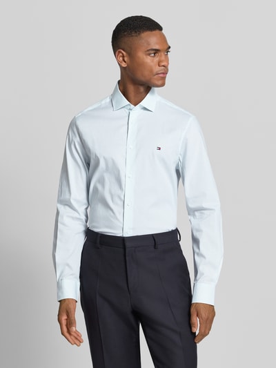 Männermodel trägt Slim Fit Businesshemd aus Baumwoll-Mix von Tommy Hilfiger, efekt: wzór w paski, wyhaftowane logo, mieszanka bawełny, koszula z kołnierzykiem typu kent, listwa guzikowa na całej długości, długi rękaw z mankietami zapinanymi na 1 guzik, regulacja szerokości, jasnoniebieski kolor, gładka tekstura, Ganzkörperansicht von vorne.