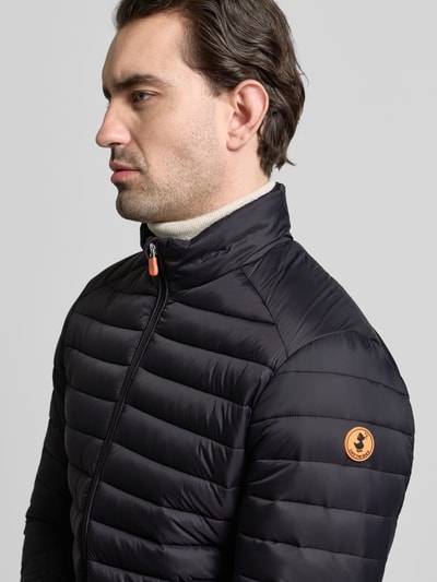 Männermodel trägt Steppjacke mit Zweiwegereißverschluss Modell 'Alexander' von SAVE THE DUCK in Schwarz, Nahaufnahme Oberkörper, Regular Fit, Stehkragen, sichtbare Markenapplikation.