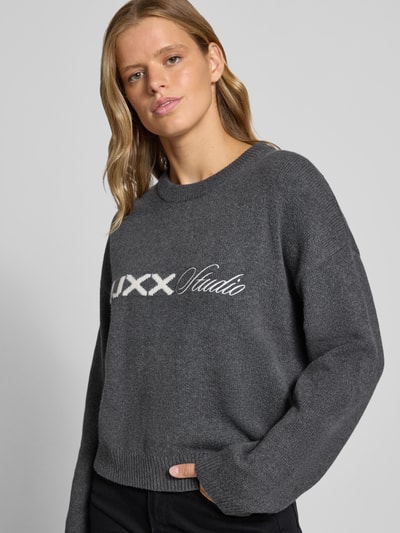Vrouwenmodel draagt Strickpullover mit Rundhalsausschnitt van JJXX, relaxed fit van katoenmix met stretch, donkergrijs met logo op het voorpand, ronde halslijn, verlaagde schoudernaden, bovenlichaam close-up.