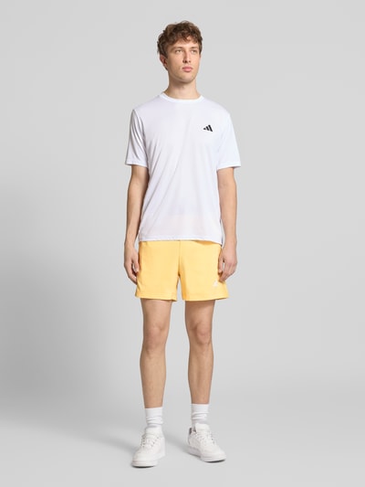 Męski model nosi Regular Fit Sport-Shorts mit Label Print od adidas Training, efekt: jednolity kolor, krój regular fit, nadruk z logo, elastyczny ściągacz z tunelem, kieszenie zapinane na zamek błyskawiczny, całkowity widok z przodu.