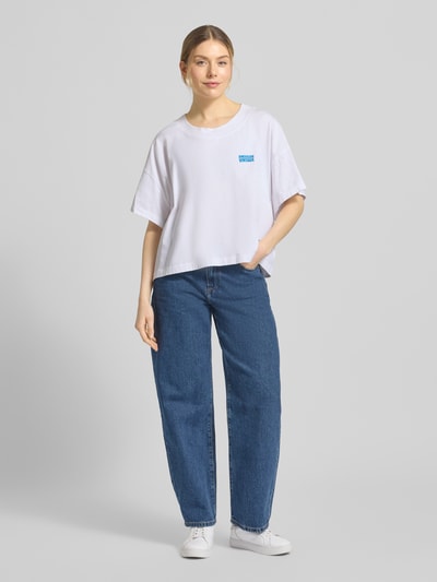 Frauenmodel trägt Damen T-Shirt mit überschnittenen Schultern Modell 'GIXY' von American Vintage in Weiß, Reine Baumwolle, Lockerer Passform, Rundhalsausschnitt, Logo-Print, Rückenlänge bei Größe XS/S 47 cm, Ganzkörperansicht von vorne.