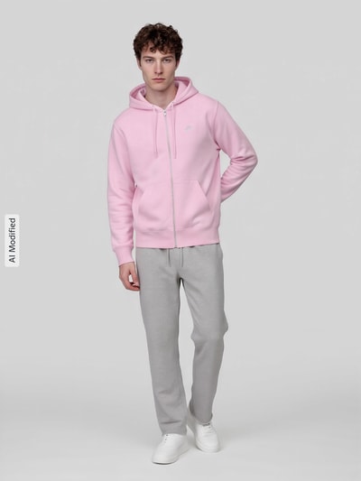 Männermodel trägt Regular Fit Sweatjacke aus Baumwoll-Mix von Nike in Rosa mit Kapuze, kombiniert mit grauer Hose, Ganzkörperansicht von vorne, KI-modifiziertes Bild.