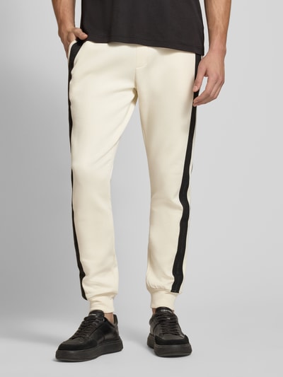 Männermodel trägt Sweatpants mit Bordüre von Armani Exchange in Creme mit schwarzem Seitenstreifen, Baumwoll-Mix, Nahaufnahme Unterkörper. Herren Sweatpants, elastischer Bund mit Tunnelzug, gerader Schnitt, Label-Print, seitliche Eingrifftaschen, Gesäßtasche, Innenbeinlänge 79 cm bei Größe M.