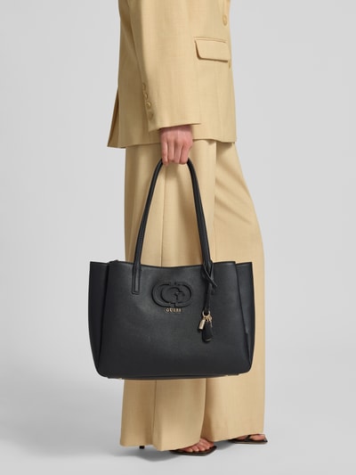 Frauenmodel trägt Tote Bag in Leder-Optik Modell 'Isola' von Guess mit Logo-Detail, geteiltem Hauptfach mit Reißverschluss und schmalen Schulterriemen, schwarze fein strukturierte Oberfläche, Maße 37 cm Breite, 27 cm Länge, 15 cm Tiefe, Schulterriemen 62 cm, Ganzkörperansicht von vorne.