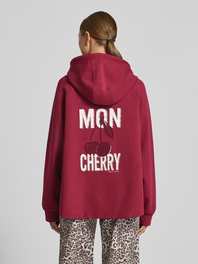 Frauenmodel trägt Relaxed Fit Hoodie mit Motiv-Print von Marc Cain in Dunkelrot, Statement-Print, Kapuze, elastische Ärmel, Ganzkörperansicht Rückansicht.