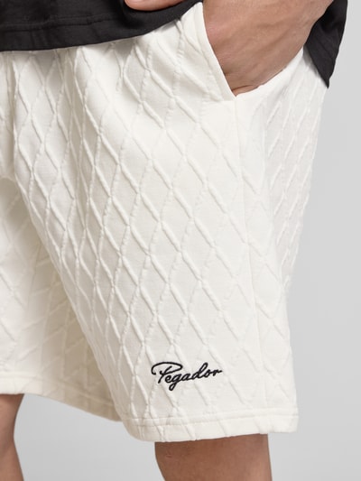 Mężczyznamodel nosi Shorts mit Logo-Stitching Modell 'Jovren' od Pegador, kremowy kolor, faktura w romby, luźny krój, detal z logo, materiał z domieszką bawełny, elastyczny pas, francuskie kieszenie, Szerokość w rozmiarze M: 38 cm, długość nogawki wewnętrznej w rozmiarze M: 24 cm, zbliżenie na górną część nogawki z przodu.