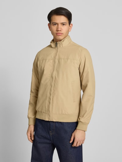 Męski model nosi Jacke mit Stehkragen und Eingrifftaschen od Superdry w kolorze beżowym, krój regular fit, zamek błyskawiczny, wpuszczane kieszenie, długość tyłu w rozmiarze M: 70 cm, szerokość ramion w rozmiarze M: 44 cm, szerokość klatki piersiowej w rozmiarze M: 41 cm, długość rękawów w rozmiarze M: 68 cm, gładka faktura, całkowity widok z przodu