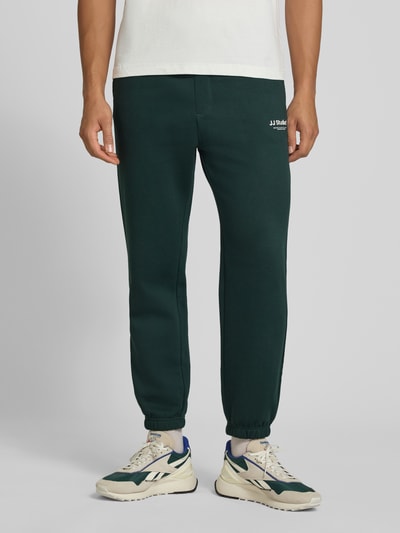 Männermodel trägt Straight Fit Sweatpants aus Baumwoll-Mix Modell 'Kane' von JACK & JONES in Dunkelgrün, Logo-Detail, elastischer Bund, elastische Beinabschlüsse, Produktkontext sichtbar, Nahaufnahme Oberkörper bis Füße.