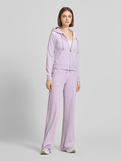 Frauenmodel trägt Slim Fit Sweatjacke aus Samt Modell 'Robertson' von Juicy Couture in Helllila, Elastischer-Material-Mix, Kapuze, Reißverschluss, geteilte Känguru-Tasche, elastische Abschlüsse, Ganzkörperansicht von vorne.