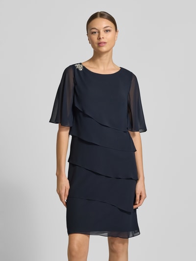 Frauenmodel trägt Relaxed Fit Kleid mit Ziersteinbesatz von Christian Berg Cocktail in Dunkelblau, semitransparent, Rundhalsausschnitt, Halbarm, Rüschen, blickdichter Unterrock, Ganzkörperansicht von vorne.
