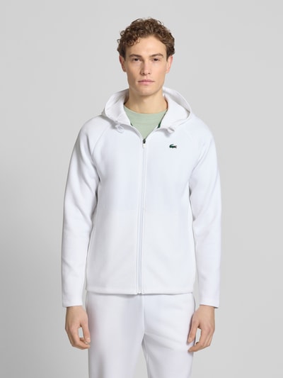 Männermodel trägt Regular Fit Sweatjacke mit Baumwoll-Anteil von Lacoste Sport in Weiß, unifarben, mit Kapuze und Logo-Patch, Ganzkörperansicht von vorne.