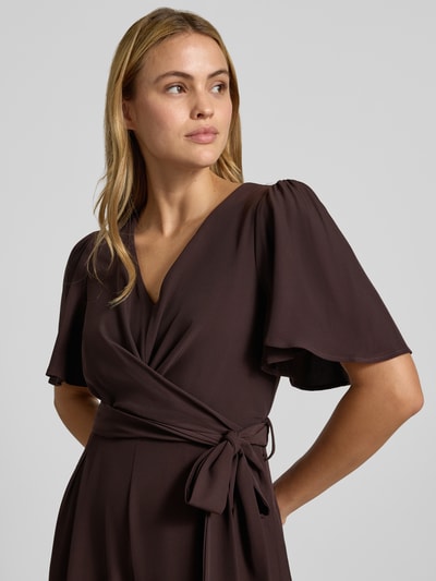 Frauenmodel trägt Jumpsuit mit Stoffgürtel von Rinascimento in Braun, V-Ausschnitt, Kurzarm, glatte Textur, Nahaufnahme Oberkörper. Damen Jumpsuit von Rinascimento, Material-Mix mit Stretch-Anteil, taillierte Passform, Raffungen, Stoffgürtel, Rückenlänge bei Größe S 148 cm, Brustweite 37 cm, Ärmellänge 30 cm, Gesamtlänge 147 cm, Innenbeinlänge 79 cm.
