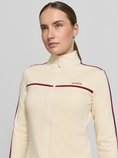 Frauenmodel trägt Slim Fit Sweatjacke mit Stehkragen Modell 'Jasmine' von Guess, cremefarben mit bordeauxroten Akzenten, Logo-Print, Nahaufnahme Oberkörper.