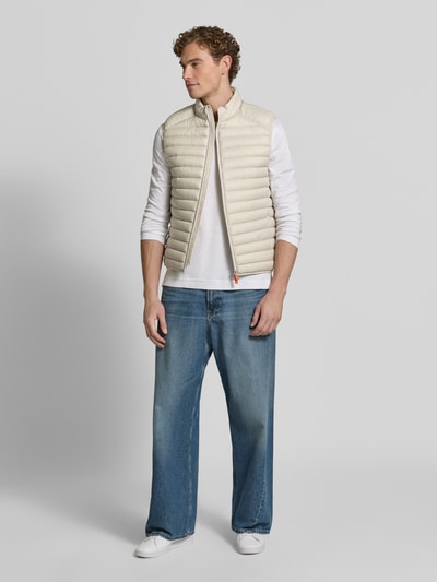Männermodel trägt Steppweste mit Zweiwege-Reißverschluss Modell 'ADAM' von Save The Duck in Beige, glatte Struktur, Ganzkörperansicht von vorne, Regular Fit, Stehkragen, Rückenlänge 61 cm, Schulterbreite 45 cm, Brustweite 57 cm.