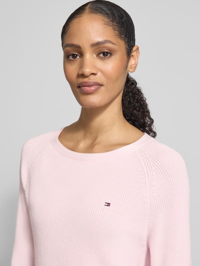 Vrouwenmodel draagt Regular Fit Pullover aus reiner Baumwolle van Tommy Hilfiger in effen lichtroze ribbreisel met logo-stiksel, ronde hals, lange mouw; close-up bovenlichaam.