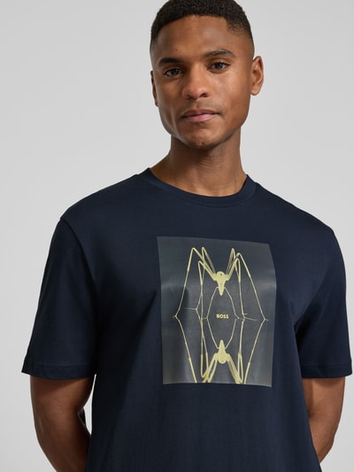 Männermodel trägt Regular Fit T-Shirt aus reiner Baumwolle Modell 'TE_CREATURE' von BOSS Orange in Dunkelblau mit gelbem Motiv-Print, Nahaufnahme Oberkörper.