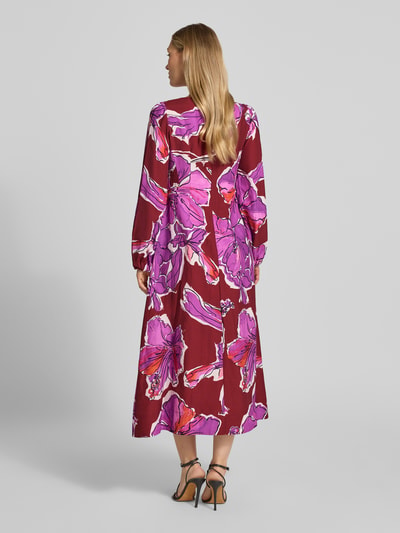 Frauenmodel trägt Midikleid aus Viskose-Mix mit V-Ausschnitt und Eingrifftaschen von Rich & Royal in Rot-Lila mit Blumenmuster, taillierter Schnitt, lange Ärmel, fließende Textur, Rückseitiger Haken- und Reißverschluss, Ganzkörperansicht Rückansicht.