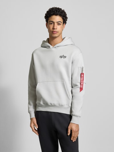 Männermodel trägt Hoodie mit Label-Details von Alpha Industries aus Baumwoll-Mix, Lockerer-Schnitt, Kapuze, ganzkörperansicht von vorne.