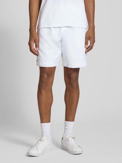 Männermodel trägt Relaxed Fit Shorts mit Logo-Patch von Lacoste in Weiß, glatte Textur, Herren Sportshorts, Ganzkörperansicht von vorne.