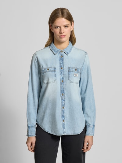 Frauenmodel trägt Regular Fit Jeansbluse mit Brusttaschen und Label-Detail von Guess aus reiner Baumwolle, Hemdblusenkragen, Knopfleiste, hellblaue Jeans-Optik, Ganzkörperansicht von vorne.