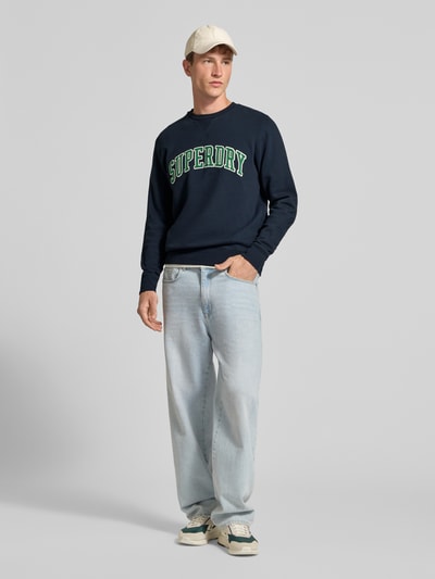 Männermodel trägt Regular Fit Sweatshirt aus Baumwoll-Mix mit Logo-Stickerei von Superdry, dunkelblau, Jeans, Ganzkörperansicht von vorne