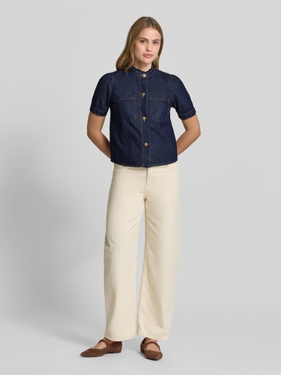 Kobiecy model nosi Regular Fit Jeansbluse mit Brusttaschen od More & More, czysta bawełna, krój regular fit, stójka, listwa guzikowa, kieszenie na piersi, długość tyłu 52 cm, długość rękawa 25 cm, ciemnoniebieski dżins, kremowe szerokie spodnie, brązowe buty, pełny widok z przodu.