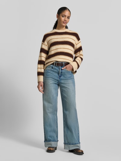 Frauenmodel trägt Strickpullover mit gerippten Abschlüssen von Mango im Material-Mix mit Baumwoll-Anteil, lockerer Schnitt, Rundhalsausschnitt, beige-braune Streifen, kombiniert mit weiter Jeans, Ganzkörperansicht von vorne.