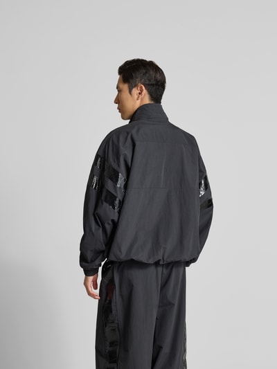 Männermodel trägt Trainingsjacke mit Stehkragen von VETEMENTS in Schwarz mit glänzenden Streifen auf den Ärmeln, Ganzkörperansicht Rückansicht, Relaxed Fit, Zweiwege-Reißverschluss, elastische Abschlüsse, Eingrifftaschen, Rückenlänge 74 cm, Schulterbreite 50 cm, Ärmellänge 73 cm.