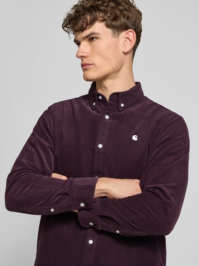 Męski model nosi Regular Fit Freizeithemd aus Cord mit Button-Down-Kragen od Carhartt Work In Progress, ciemnofioletowa bawełna, label-stitching, krój regular fit, model Madison, długość tyłu w rozmiarze M 76 cm, szerokość ramion w rozmiarze M 44 cm, długość rękawa w rozmiarze M 64 cm, zbliżenie na górną część ciała z przodu