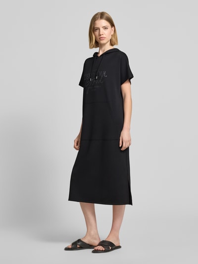 Frauenmodel trägt Sweatkleid mit Kapuze von Soyaconcept in Schwarz, Material-Mix mit Modal-Anteil, Gerader-Schnitt, Midikleid, Label-Print. Ganzkörperansicht von vorne.