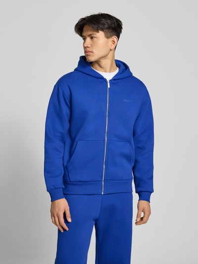 Männermodel trägt Sweatjacke mit Känguru-Tasche von REVIEW in Blau aus Baumwoll-Mix, Relaxed-Fit, Reißverschluss, Ganzkörperansicht von vorne. Rückenlänge bei Größe M: 68 cm, Ärmellänge bei Größe M: 62 cm.