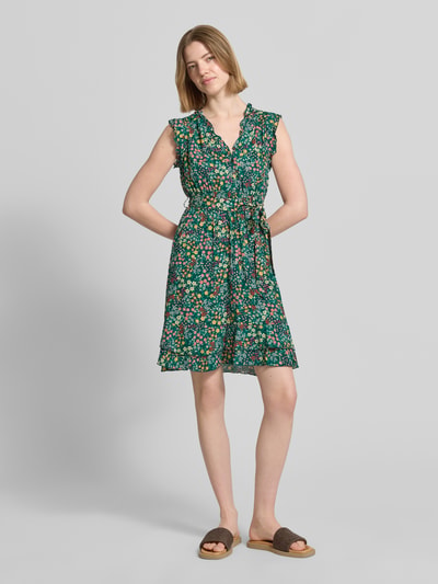 Frauenmodel trägt Minikleid aus reiner Viskose mit floralem Allover-Print von Apricot, taillierte Passform, Rüschenbesatz, abnehmbares Taillenband, Ärmelloses-Design, V-Ausschnitt, Knopfleiste, grüne Grundfarbe mit buntem Blumenmuster, Rückenlänge bei Größe S: 85 cm, Brustweite 30 cm, Ärmellänge 6 cm, Ganzkörperansicht von vorne.