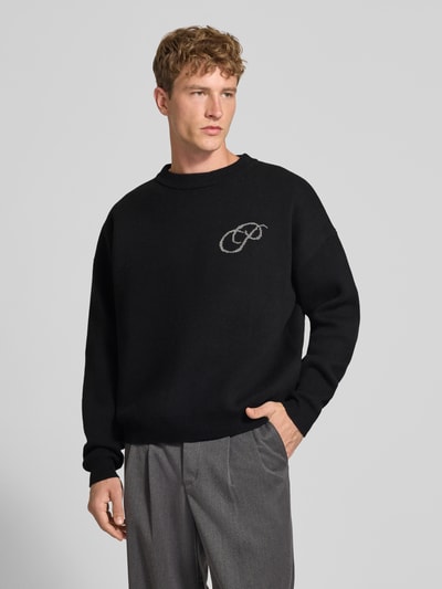 Męski model nosi Relaxed Fit sweter z dzianiny z bawełny od PROHIBITED, wyhaftowane logo, prążkowane wykończenia, Długość tyłu w rozmiarze M: 64 cm, Długość rękawów w rozmiarze M: 59 cm, luźny krój, całość w kolorze czarnym, widok całej sylwetki z przodu.