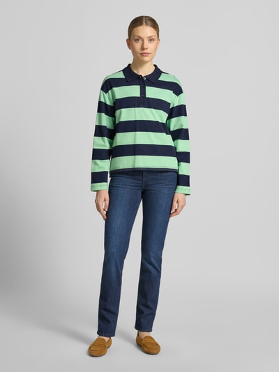 Modelka kobieca nosi Loose Fit Poloshirt aus reiner Baumwolle od s.Oliver RED LABEL, zielono-granatowe pasy, prążkowany materiał, długi rękaw, przód z krótką listwą guzikową, kołnierz polo, przód z rozcięciami po bokach, całość z bawełny, widok całopostaciowy z przodu.