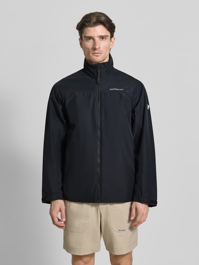 Mężczyzna nosi Regular Fit Jacke mit Logo-Print Modell 'Quest' od PEAK PERFORMANCE, czarna, stojący kołnierz, kieszenie, zamek, długość tyłu w rozmiarze M: 74 cm, szerokość ramion w rozmiarze M: 45 cm, szerokość klatki piersiowej w rozmiarze M: 55 cm, długość rękawa w rozmiarze M: 65 cm, widok całej sylwetki z przodu.