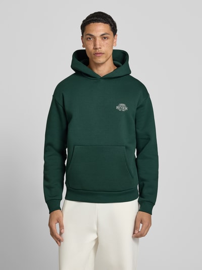 Männermodel trägt Hoodie mit Logo-Print und Känguru-Tasche von REVIEW in Dunkelgrün, Baumwoll-Mix, Relaxed-Fit, Gerippte-Abschlüsse, Ganzkörperansicht von vorne.