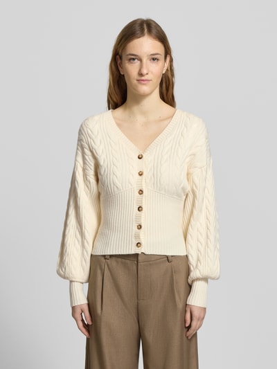 Modelka nosi Strickjacke mit Knopfleiste od Gina Tricot, kremowy kolor, krój regular fit, wiskoza, prążkowane wykończenia, dekolt w serek, dł. tyłu 48 cm, dł. rękawa 52 cm, ujęcie całej sylwetki z przodu.