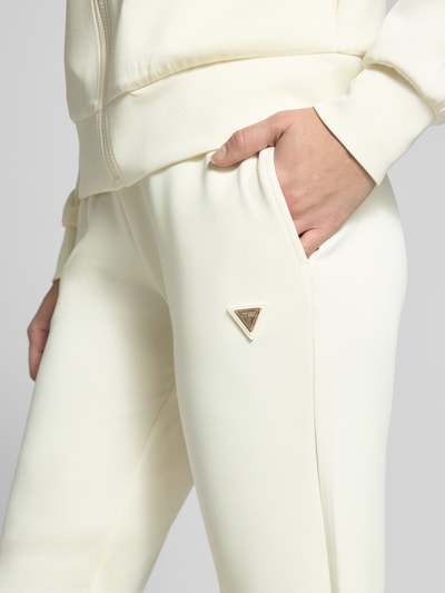 Kobieta nosi Sweatpants mit Label-Detail od Guess Activewear, kremowy kolor, elastyczny materiał z domieszką modalu, prosta nogawka, elastyczny pas, kieszenie boczne, widoczne logo, zbliżenie na biodra.