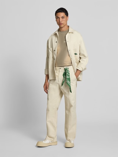 Männermodel trägt Straight Fit Hose aus Reiner Baumwolle von Forét in Beige mit Eingrifftaschen, Gesäßtaschen, Knopf-Reißverschluss, Ganzkörperansicht von vorne.