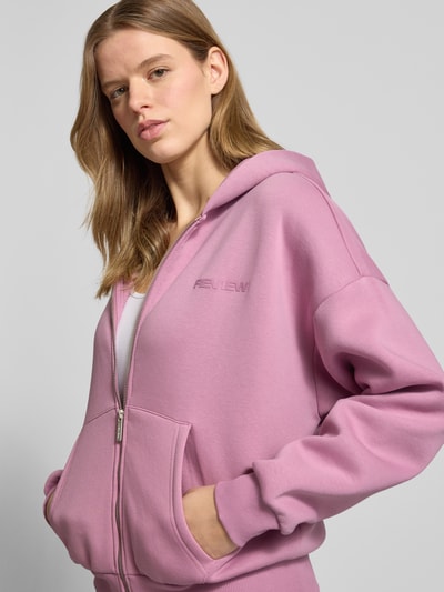 Frauenmodel trägt Sweatjacke mit Logo und Känguru-Tasche von Review in Rosa, Baumwoll-Mix, Relaxed-Fit, Kapuze, Reißverschluss, Ganzkörperansicht von vorne.