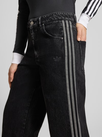 Kobiece model prezentuje 'Loose Fit Jeans aus reiner Baumwolle' od adidas Originals, denim, krój loose fit, naszywka z logo, paski w kontrastowym kolorze, zapięcie na guzik i zakryty zamek błyskawiczny, 5 kieszeni, czysta bawełna, zbliżenie na górną część nogawki