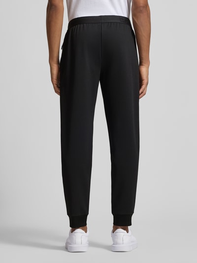 Männermodel trägt zwarte Sweatpants mit elastische band und labelpatch van EA7 Emporio Armani, materiaalmix met viscose, relaxed fit, binnenbeenlengte maat M 77 cm, ganzkörperansicht rückansicht.