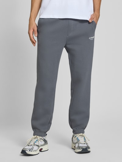 Männermodel trägt Straight Fit Sweatpants aus Baumwoll-Mix Modell 'Kane' von JACK & JONES in Grau mit Logo-Detail, Nahaufnahme Unterkörper. Herren Sweatpants aus Baumwoll-Mix mit Gesäßtasche, elastischem Bund, elastischen Beinabschlüssen, Innenbeinlänge 72 cm bei Größe M.