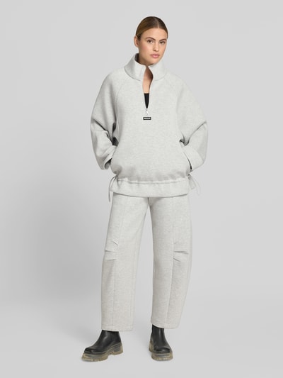 Frauenmodel trägt Sweatshirt aus Viskose-Mix von ROTATE in Hellgrau mit Logo-Patch, Stehkragen, Reißverschluss und taillierter Passform, Ganzkörperansicht von vorne.
