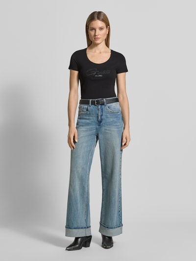 Frauenmodel trägt T-Shirt mit U-Ausschnitt Modell 'SCRIPT' von Guess aus Baumwoll-Elasthan-Mix, körpernaher Schnitt, Label-Stitching, schwarze Farbe, Rückenlänge bei Größe S 61 cm, Ganzkörperansicht von vorne.