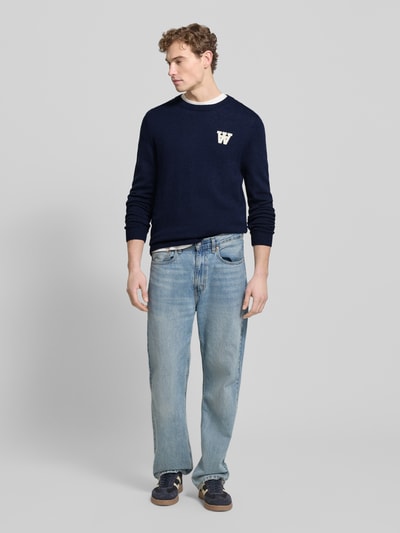 Männermodel trägt dunkelblauen Strickpullover aus Lammwolle Modell 'Tay' von Wood Wood, Regular-Fit, Rundhalsausschnitt, Label-Stitching, Ganzkörperansicht von vorne.
