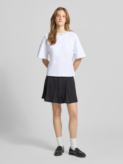 Frauenmodel trägt weißes Comfort Fit T-Shirt mit Label-Schriftzug von Calvin Klein Jeans, kombiniert mit schwarzem Rock; Ganzkörperansicht von vorne.