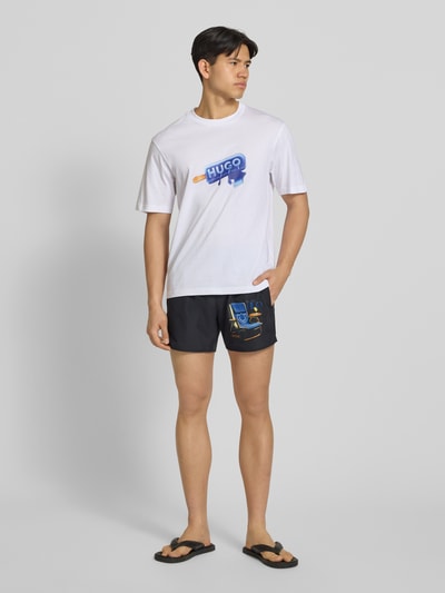 Männermodel trägt Badeshorts mit motiefprint van Hugo Blue in effen kleuren met achterzak en steekzakken, materiaalverwerking popeline. Ganzkörperansicht von vorne.