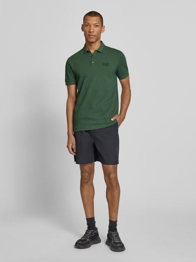 Męski model nosi Regular Fit Poloshirt mit Logo-Print od EA7 Emporio Armani, bawełna elastyczna, krótki rękaw, kolor zielony, widok całej sylwetki z przodu.